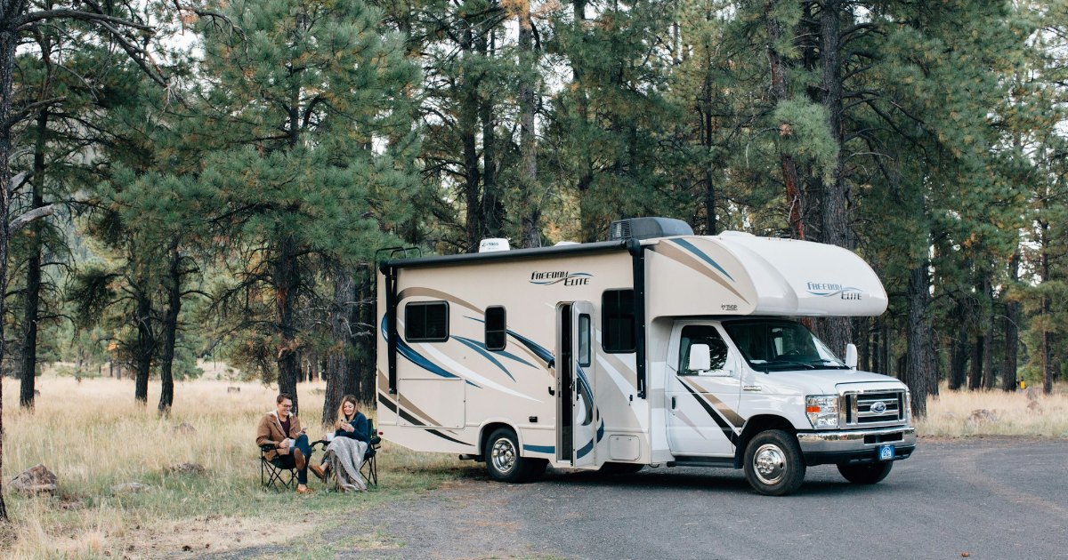 A couple relaxing in camp chairs beside a Class C motorhome at a forest campsite